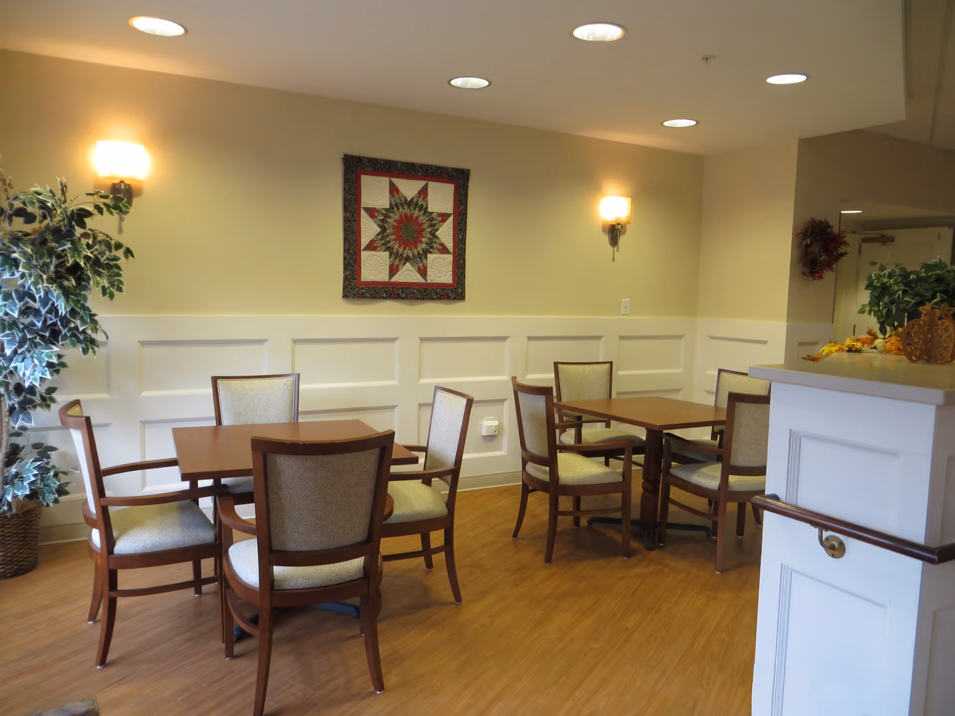 Small communal dining area with wooden tables and upholstered chairs, wall sconces, a decorative quilt on the wall, and potted plants.