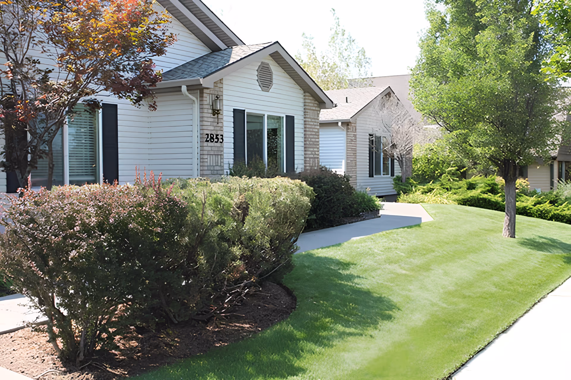 Exterior view of a single-story residential building with white siding and brick accents, surrounded by well-maintained green grass, bushes, and trees under a sunny sky.