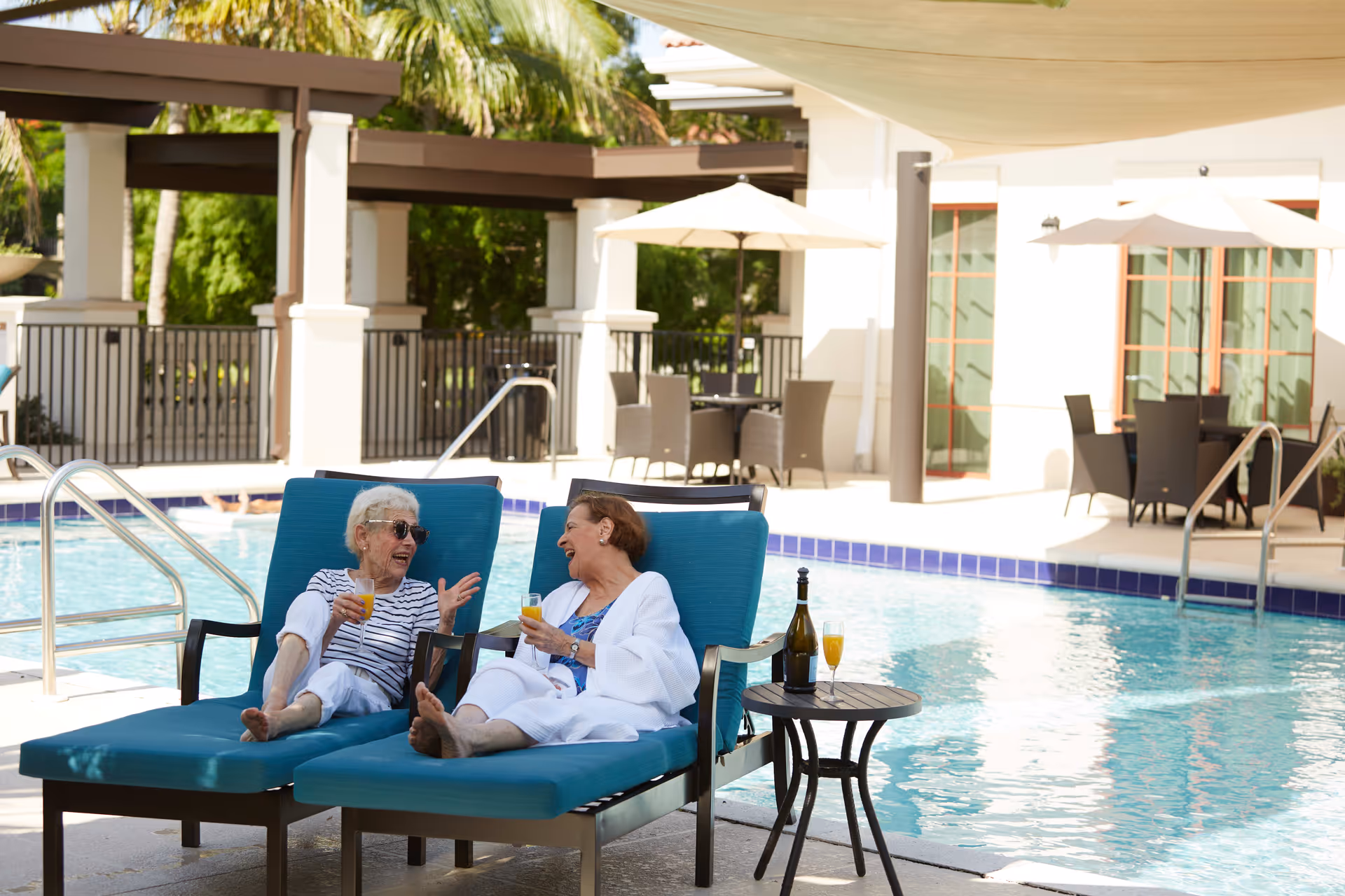 Two elderly women relaxing and chatting on blue lounge chairs by a swimming pool at an outdoor patio area. They are holding glasses of orange juice, with a bottle and two more glasses on a small table beside them. The background shows a building with large windows, patio tables with umbrellas, and palm trees.