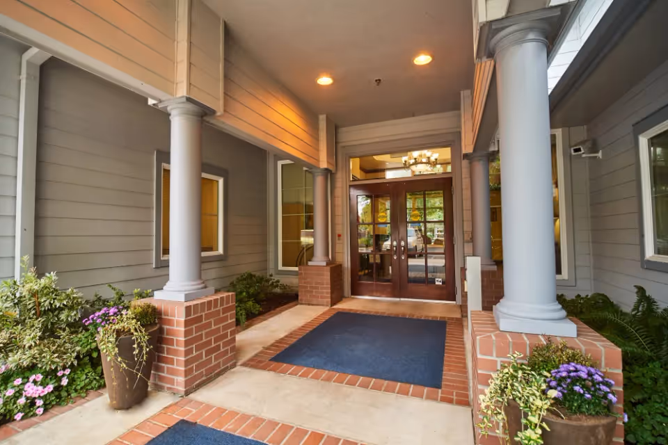 Entrance to a building with double glass doors framed by brick and white columns, flanked by potted plants and greenery under a covered porch with ceiling lights.