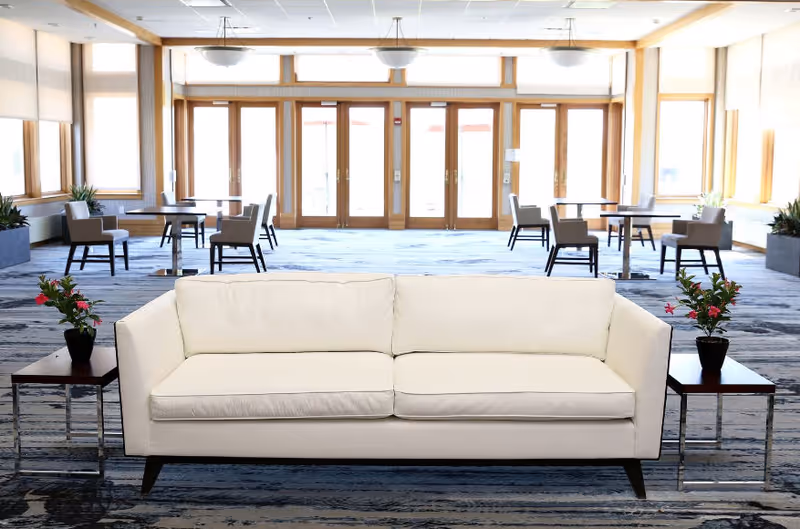 A bright and spacious common area with a white sofa centered in the foreground, flanked by two small tables each holding a potted plant with red flowers. In the background, there are multiple tables and chairs arranged near large windows and glass doors that let in natural light.