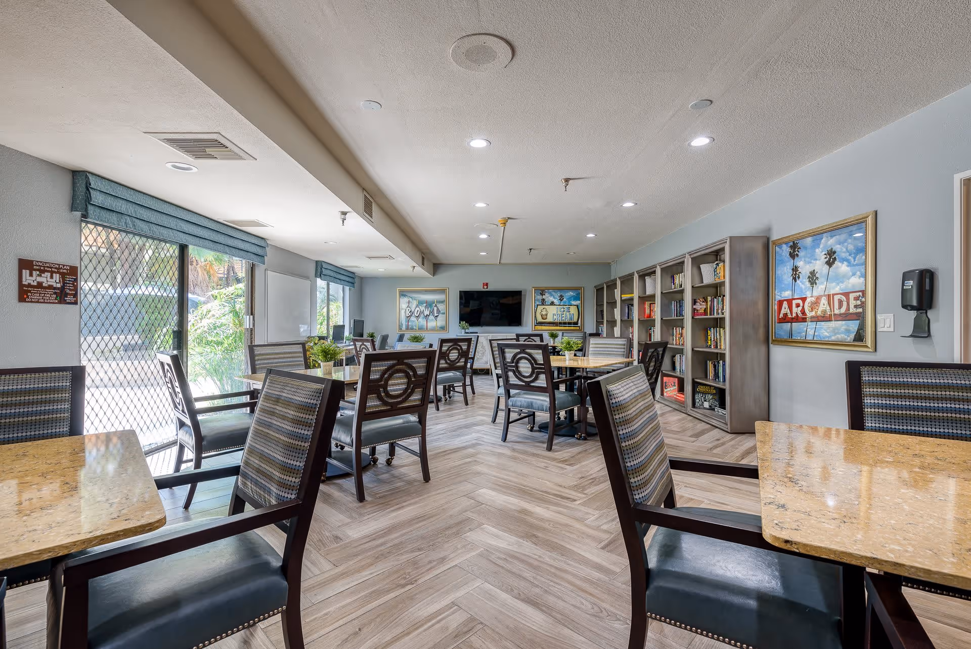 A bright and spacious common area in a senior living facility with multiple tables and chairs arranged for seating. The room features large windows with blue valances, a bookshelf filled with books and games, and framed artwork on the walls. A flat-screen TV is mounted on the far wall above a fireplace. The flooring is light wood in a herringbone pattern.