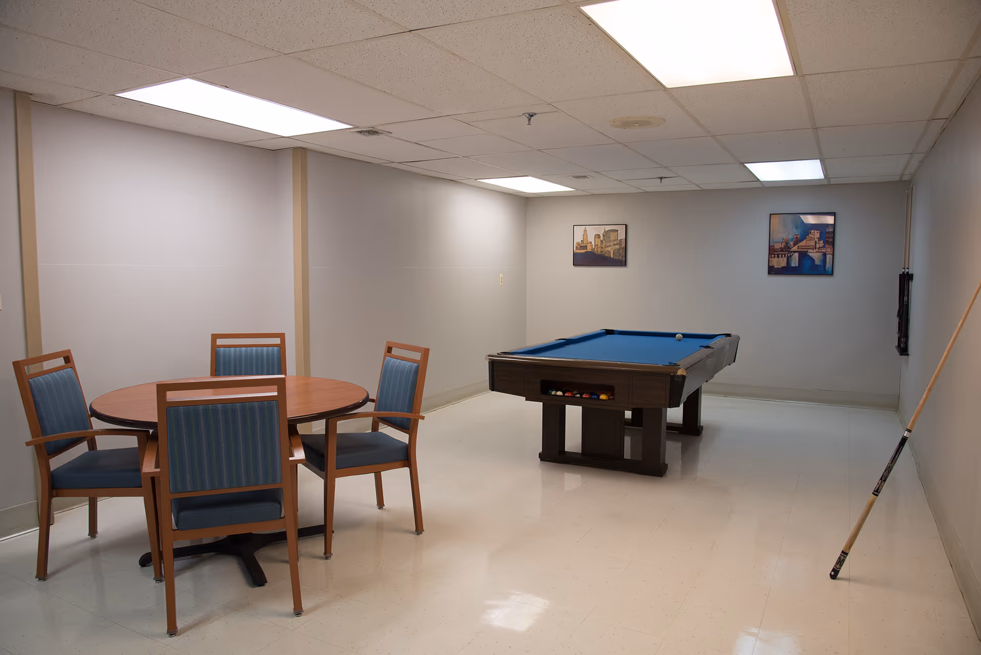 A recreational room with a pool table and a round wooden table surrounded by four chairs with blue cushions. The room has plain light-colored walls, a tiled floor, and fluorescent ceiling lights. Two framed pictures hang on the far wall, and a pool cue is leaning against the right wall.