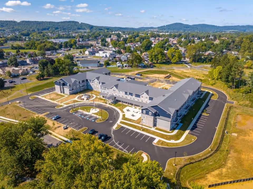 Aerial view of The Residence at Village Greens, a large senior living facility with multiple connected buildings, surrounded by parking lots and green spaces, set against a backdrop of a small town and rolling hills under a partly cloudy sky.