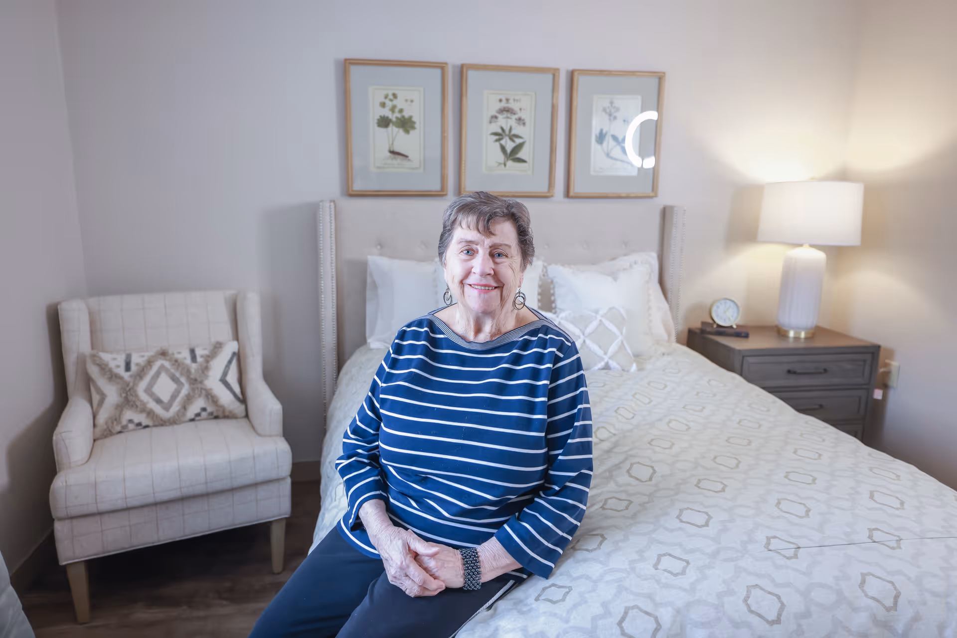 An elderly woman wearing a blue and white striped shirt sits on the edge of a neatly made bed in a cozy bedroom. Behind her, there are three framed botanical prints on the wall. To the left of the bed is a cushioned armchair with a decorative pillow, and to the right is a nightstand with a lamp and a small clock.