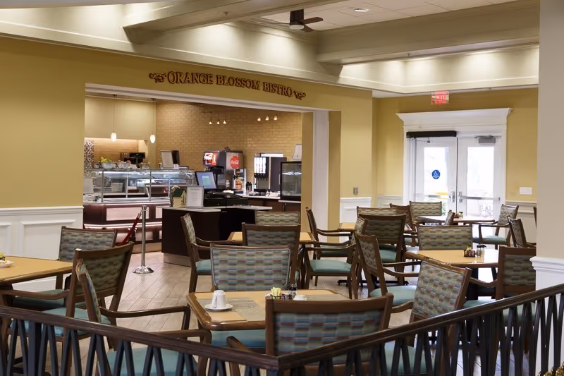 Interior view of a dining area named Orange Blossom Bistro with multiple tables and chairs arranged neatly. The room has soft yellow walls, white trim, and a counter area with beverage dispensers and food service equipment in the background. There is a double glass door exit on the right side.