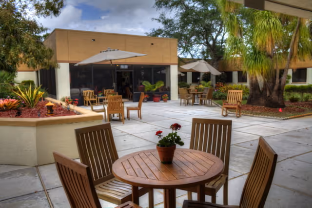 Outdoor patio area with multiple wooden tables and chairs, some tables shaded by large umbrellas. The patio is surrounded by landscaped garden beds with plants and trees, and a building with large windows is visible in the background under a partly cloudy sky.