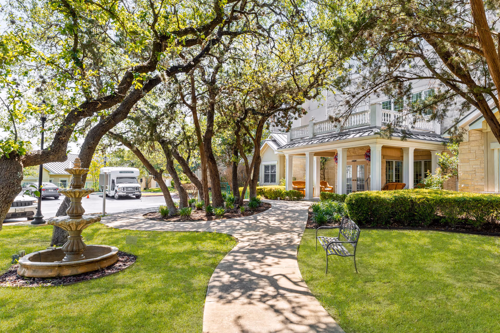 A sunny outdoor pathway lined with trees and greenery leads to the entrance of a building with a covered porch. There is a three-tiered stone fountain on the left side of the path and a metal bench on the right side. Several cars and a white shuttle bus are visible in the background near the parking area.