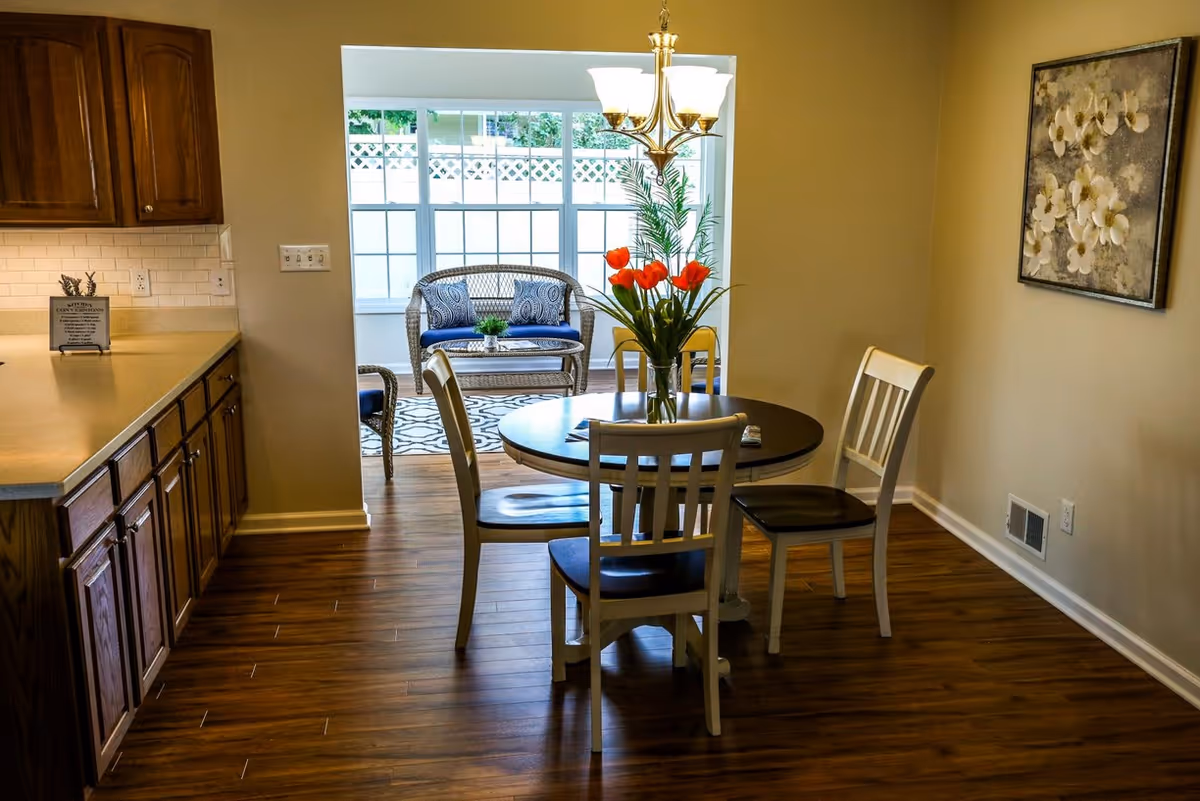 A dining area with a round wooden table and four chairs, a vase with red tulips on the table, adjacent to a kitchen with wooden cabinets and a countertop. In the background, there is a sunroom with wicker furniture and large windows letting in natural light. A floral painting hangs on the wall to the right.