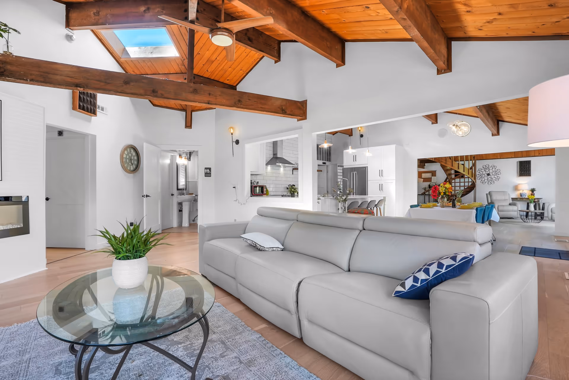 Spacious living room with a large light gray leather sofa adorned with two pillows, a glass coffee table with a potted plant, wooden ceiling beams with a skylight, and an open view into a modern kitchen and dining area with a spiral staircase in the background.