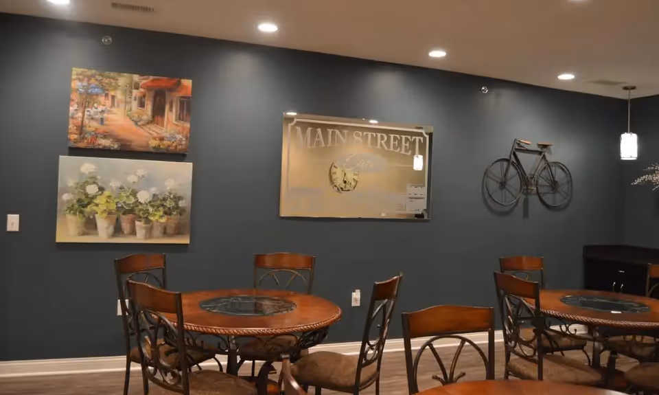 Interior view of a dining area with round wooden tables and metal chairs with cushioned seats. The walls are painted dark gray and decorated with two paintings, a mirror with the text 'MAIN STREET Cafe', and a decorative bicycle mounted on the wall. Ceiling lights illuminate the room.