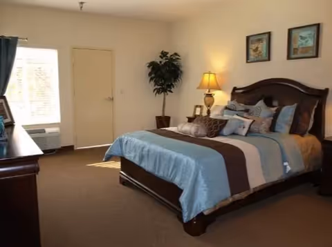 Furnished bedroom featuring a large bed with blue and brown bedding, a nightstand with lamp, dresser, window and potted plant.
