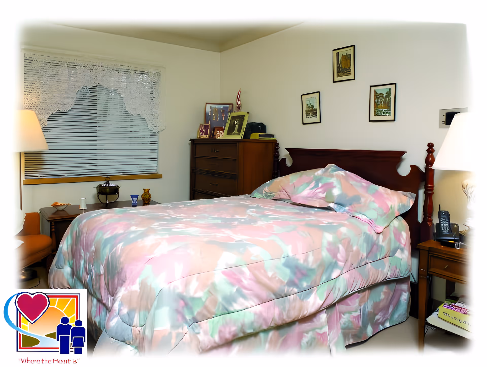 A bedroom with a bed covered in a pastel-colored floral comforter and matching pillow. The room has a wooden headboard, a wooden dresser with framed photos, a window with blinds and lace curtains, two bedside tables with lamps, and framed pictures on the wall.
