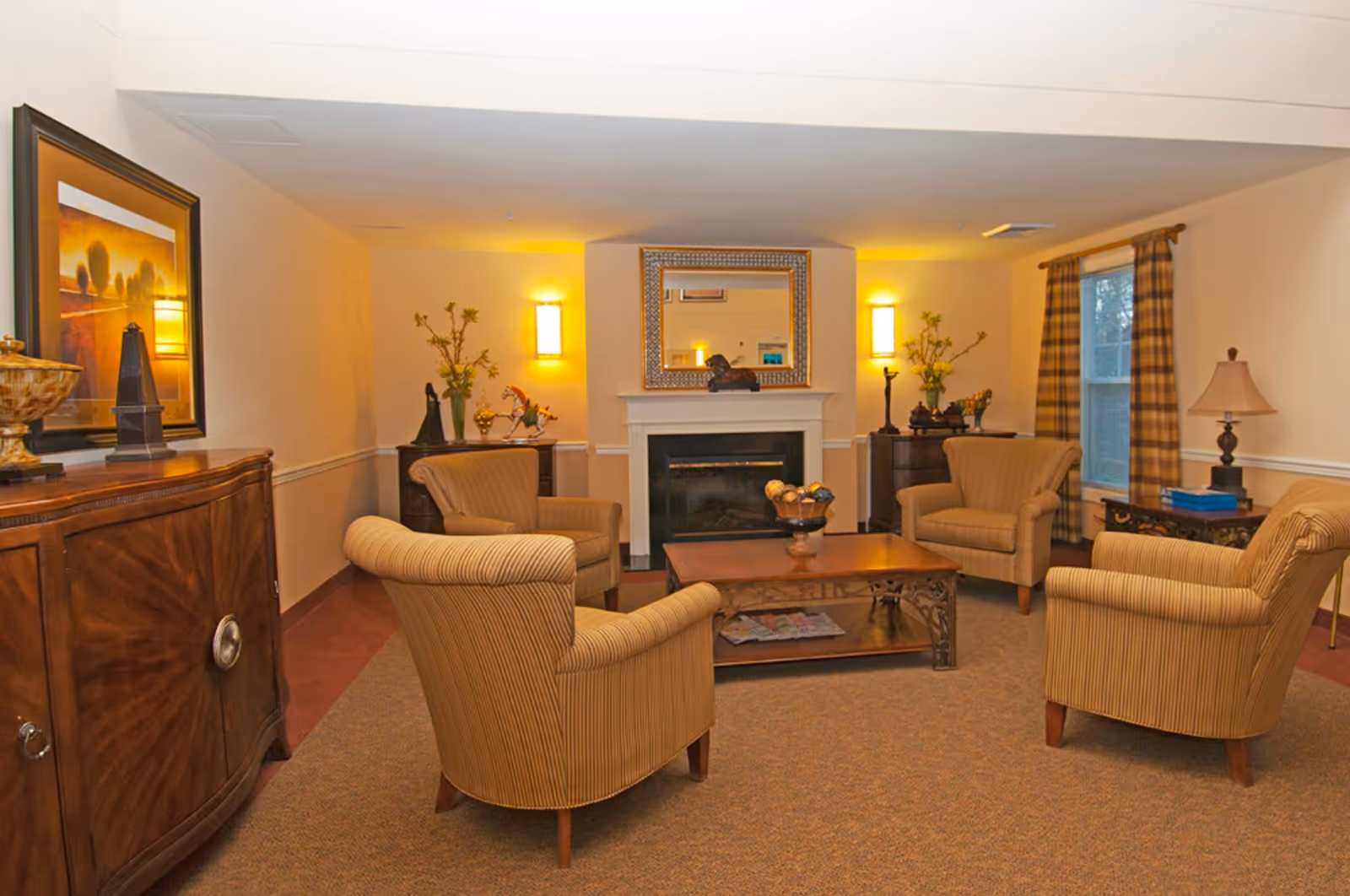 A cozy living room with four beige striped armchairs arranged around a wooden coffee table. The room features a fireplace with a decorative mirror above it, two wall sconces providing warm lighting, and two windows with plaid curtains. There are wooden side tables with lamps and decorative items, and a wooden cabinet with framed artwork on the left side.