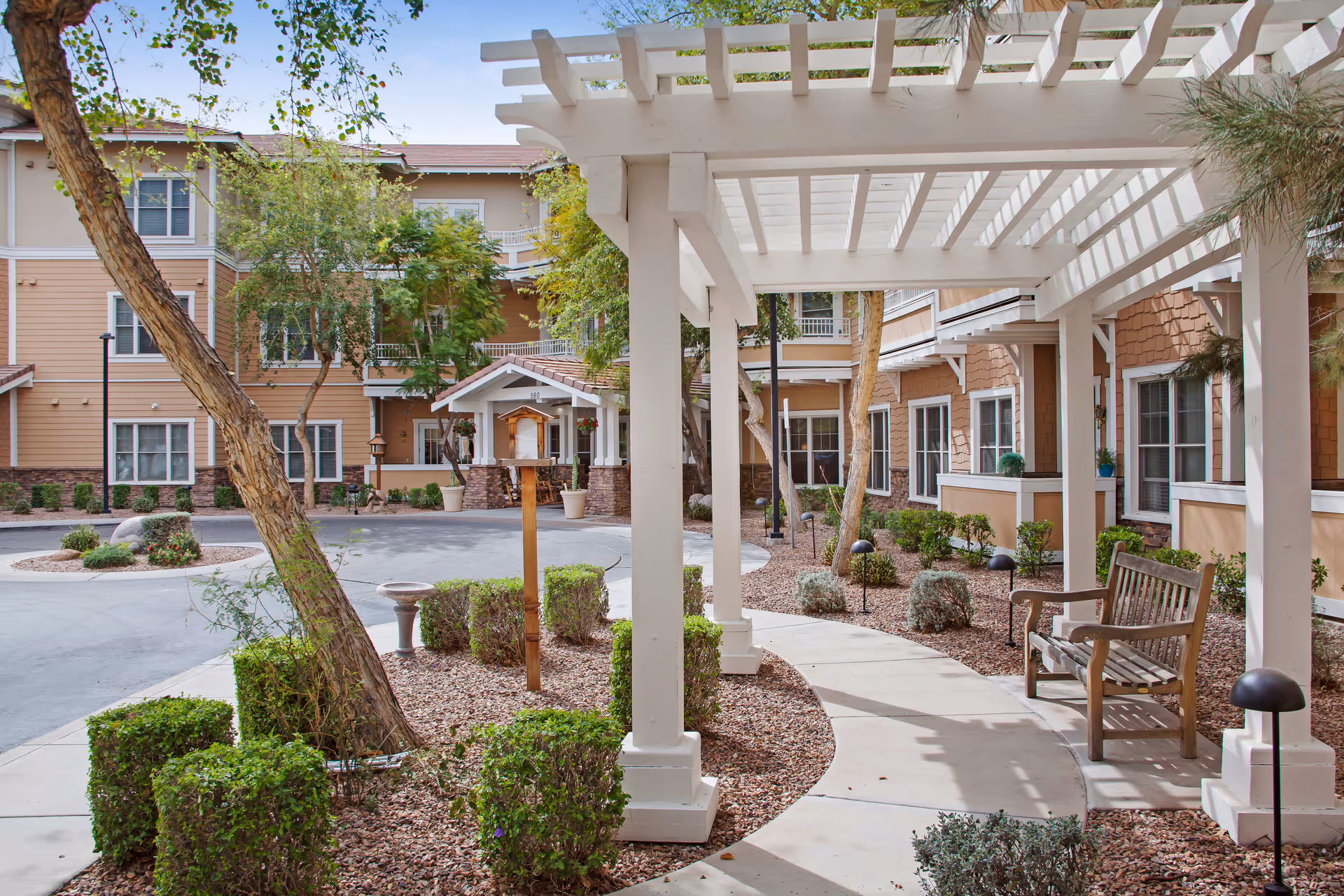 Outdoor courtyard area of a senior living facility with a white pergola, wooden bench, landscaped bushes, trees, and a curved concrete walkway leading to the building entrance.