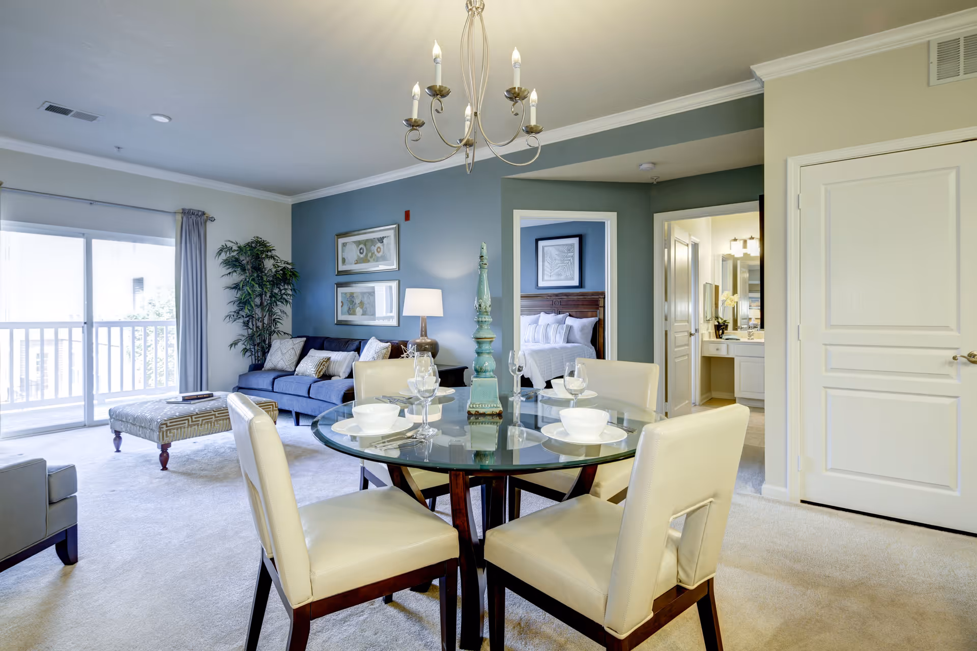 A bright and modern living and dining area in a senior living facility. The dining area features a round glass table set with white bowls, plates, and wine glasses, surrounded by four cream-colored chairs. The living room has a blue sofa with cushions, a patterned ottoman, a floor lamp, and framed artwork on a blue accent wall. A large sliding glass door with curtains leads to a balcony. In the background, a bedroom with a neatly made bed and a bathroom with a vanity and mirror are visible through open doorways.