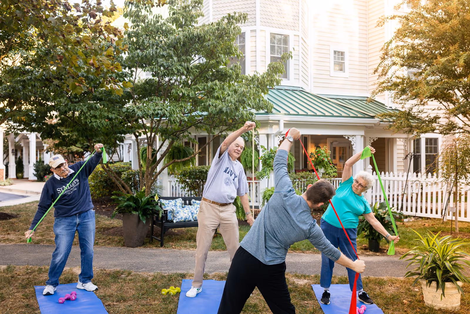 Four seniors participating in an outdoor exercise session using resistance bands on blue mats in front of a white residential building with a green roof and white picket fence. Trees and plants surround the area, creating a peaceful garden setting.