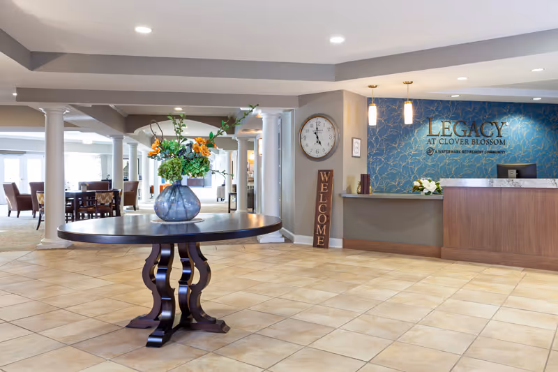 Spacious and well-lit reception area of a senior living facility with a round wooden table holding a vase of flowers in the center. To the right is a reception desk with a blue decorative wall behind it displaying the text 'LEGACY AT CLOVER BLOSSOM'. A clock and a vertical welcome sign are mounted on the wall near the desk. In the background, there are columns and seating areas with tables and chairs.