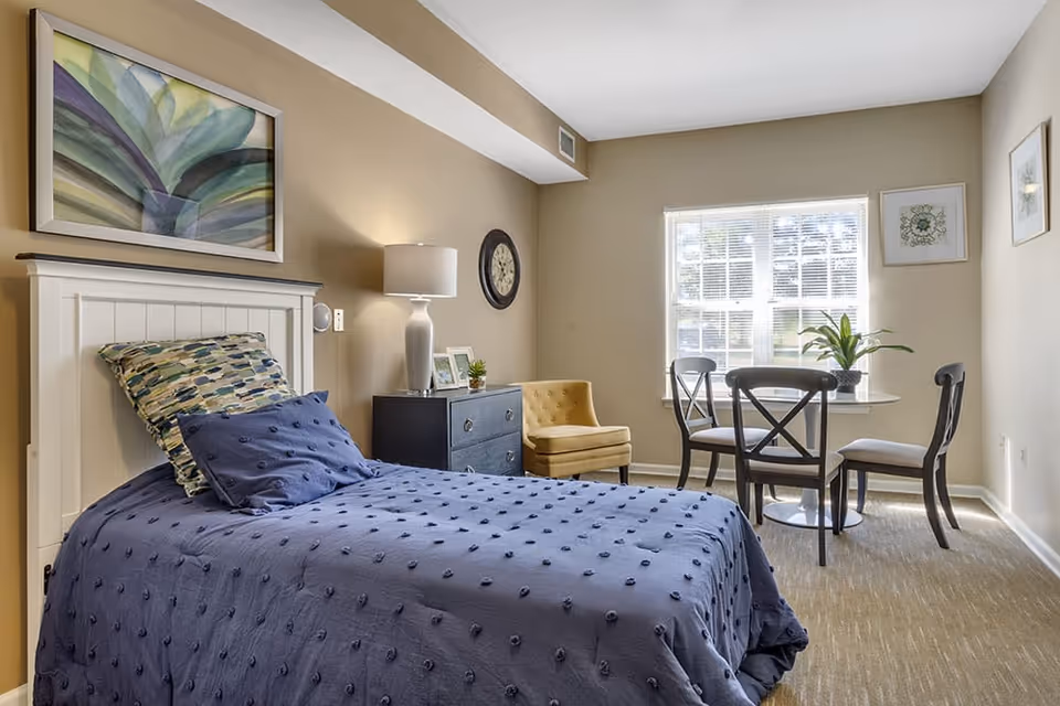 A cozy bedroom with a single bed covered in a blue textured blanket and two pillows. Next to the bed is a dark blue dresser with a white lamp, small plant, and framed photos. A yellow upholstered chair is positioned near a window with white blinds, beside a small round table with three chairs and a potted plant. The walls are beige with framed artwork and a round clock.