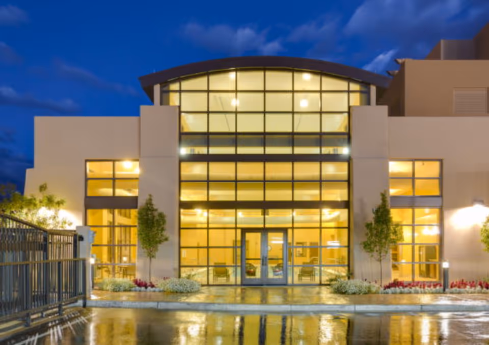 Exterior front view of a modern building at dusk with large glass windows and doors illuminated from inside, reflecting on a wet pavement. The building has a curved roof section and is surrounded by small trees and flower beds.