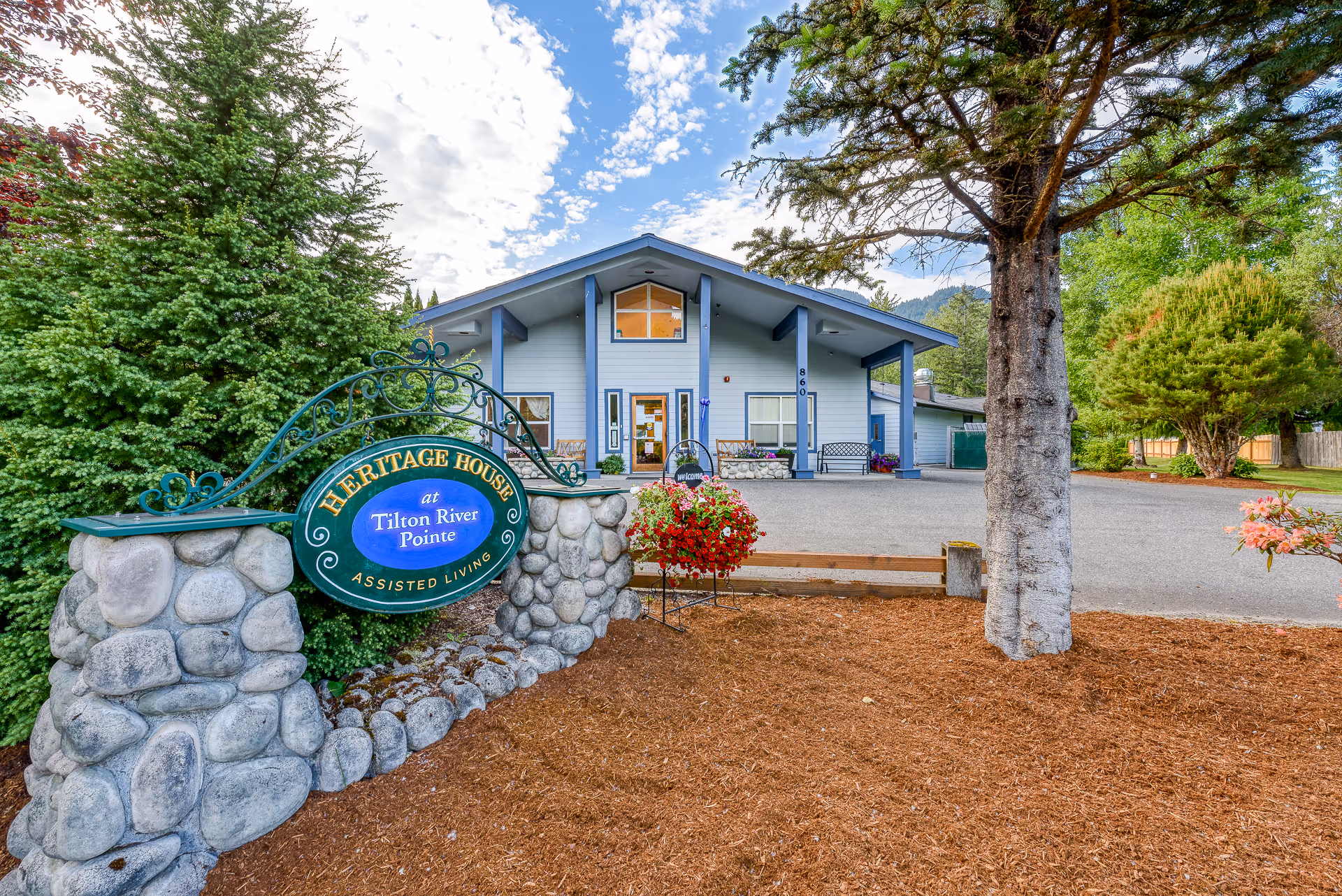 Exterior view of Heritage House at Tilton River Pointe Assisted Living facility showing a light blue building with a peaked roof, surrounded by trees and landscaping. A stone sign with green and gold lettering is visible in the foreground, along with a hanging flower basket and a large tree on the right side.