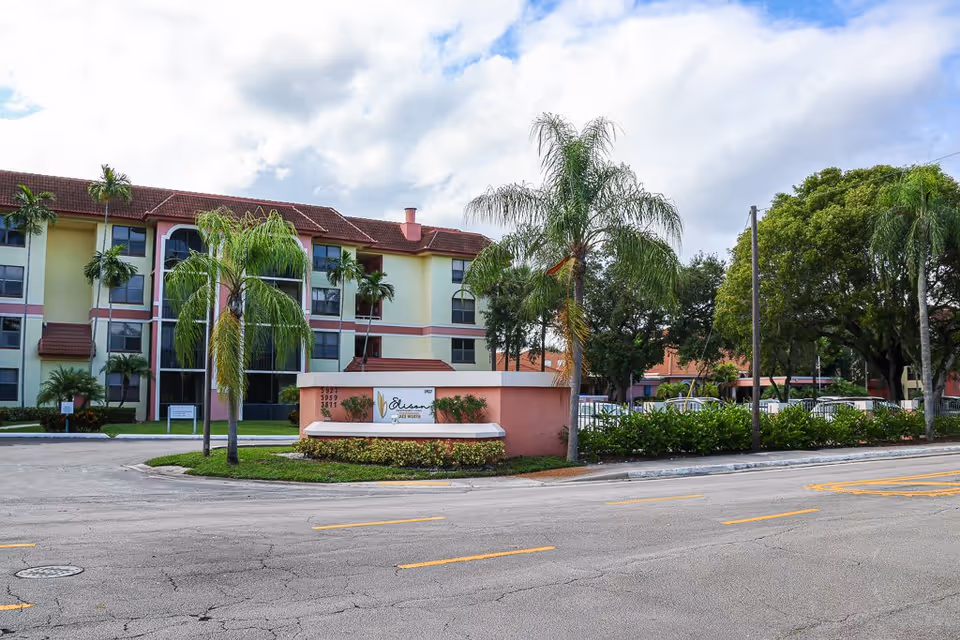 Exterior view of Elison Independent Living of Lake Worth, showing a multi-story residential building with a red-tiled roof, palm trees, and a sign with the facility's name near the entrance along a paved road.
