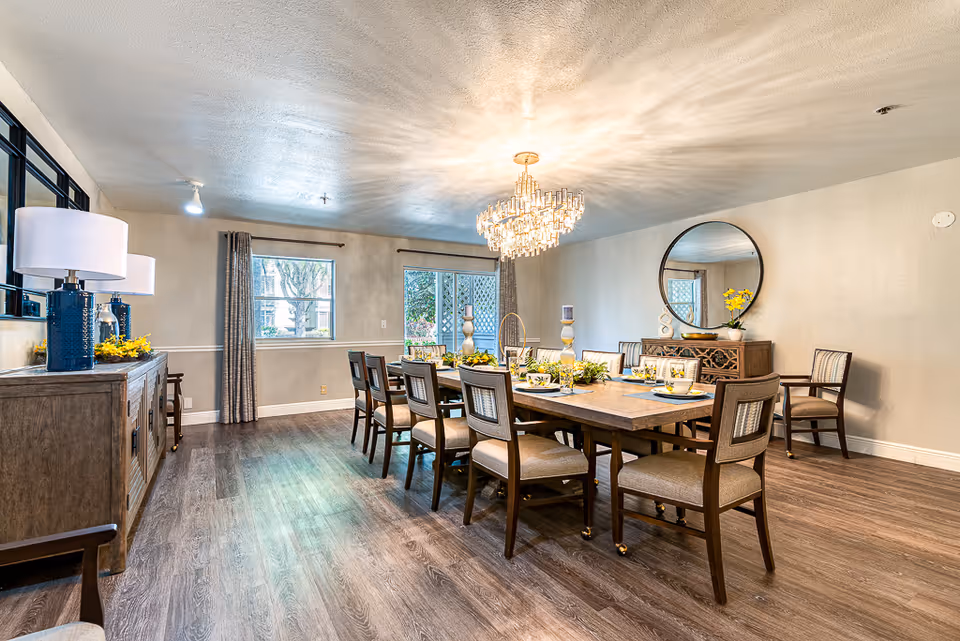 A spacious dining room with a large rectangular wooden table surrounded by ten cushioned chairs. The table is set with plates, glasses, and decorative centerpieces including candles and flowers. A modern chandelier hangs above the table, casting light across the room. The walls are light-colored with a large round mirror and a wooden sideboard decorated with a vase of yellow flowers and other ornaments. Two windows with curtains allow natural light into the room, and the floor is covered with wood-style flooring.