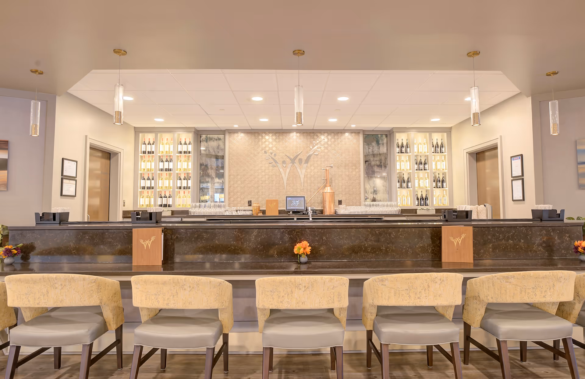 A modern bar area with six beige cushioned bar stools lined up in front of a dark marble countertop. Behind the bar, there are shelves with bottles of wine and glasses, and a decorative wall with a logo featuring two stylized human figures. The ceiling has recessed lighting and hanging pendant lights.