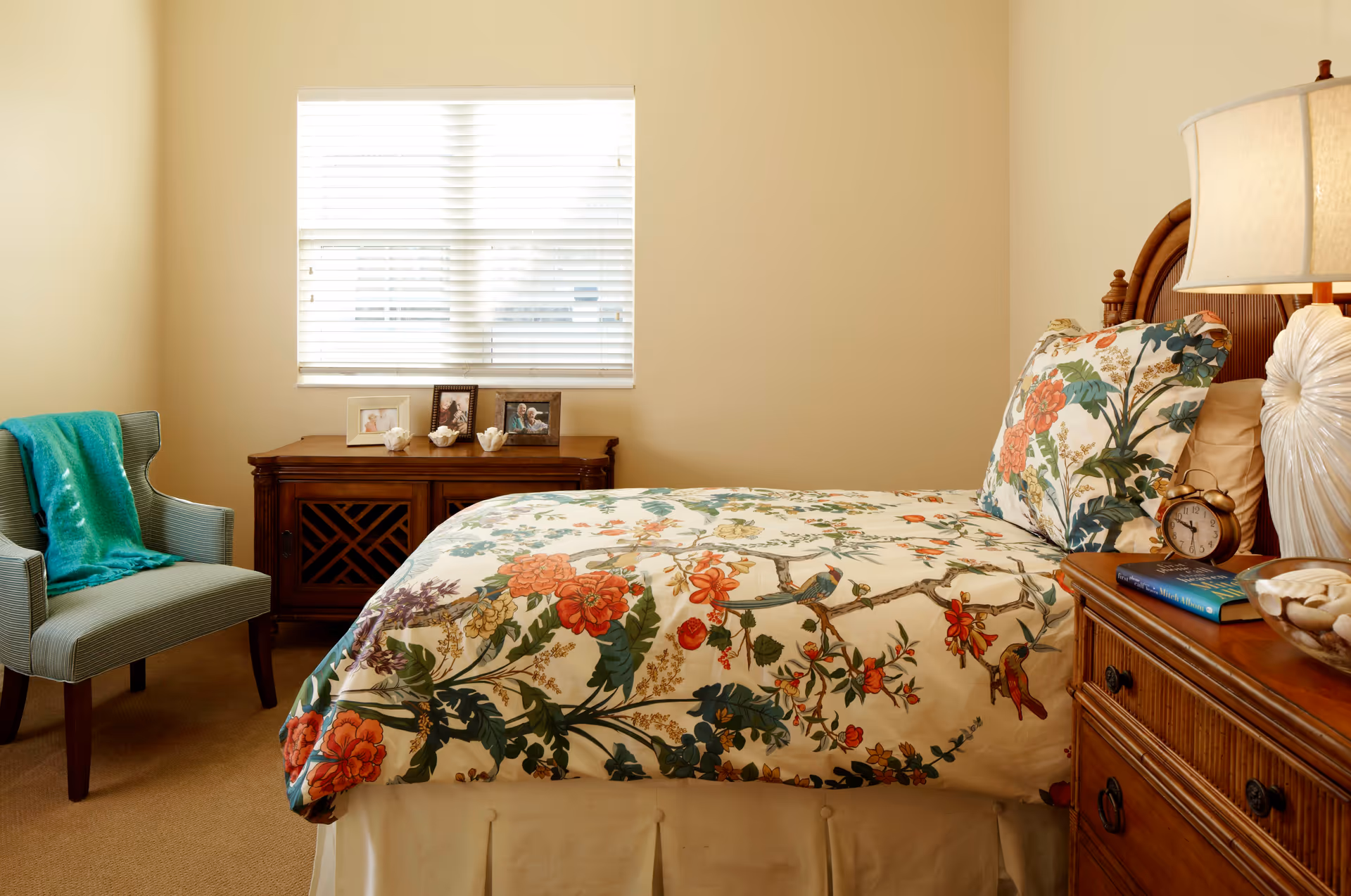 A cozy bedroom with a bed featuring floral and bird patterned bedding. To the right of the bed is a wooden nightstand with a lamp, an alarm clock, a book, and a decorative bowl. Against the back wall is a wooden cabinet with framed photos and decorative items on top. To the left is a blue upholstered chair with a teal throw draped over it. A window with white blinds lets in natural light.