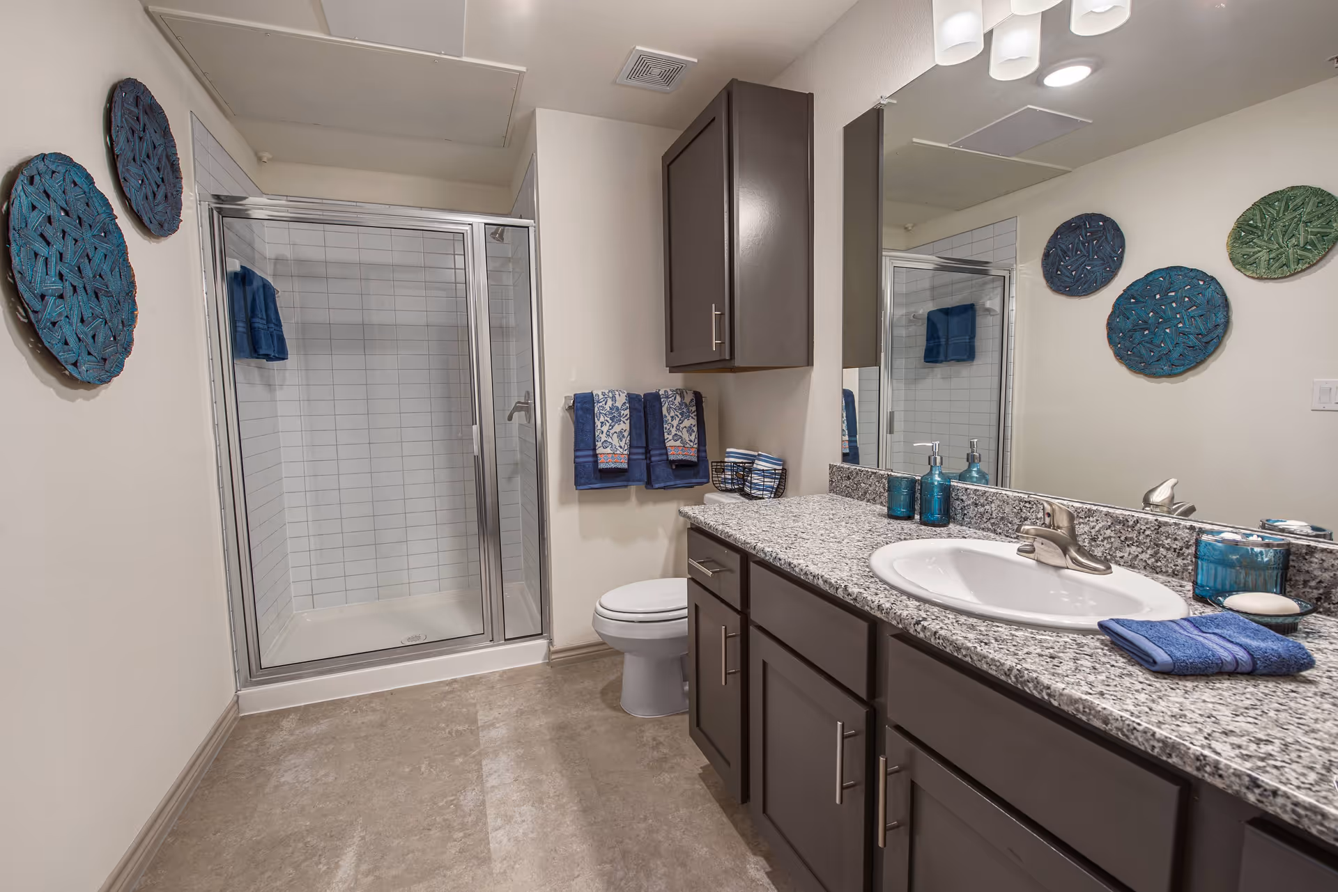 A modern bathroom featuring a glass-enclosed shower with white subway tiles, a toilet, and a granite countertop with a white sink. The vanity has dark wood cabinets and drawers. Blue and patterned towels hang on a rack next to the toilet. Decorative blue and green woven wall hangings are displayed on the walls. The bathroom has a large mirror above the sink and recessed lighting in the ceiling.