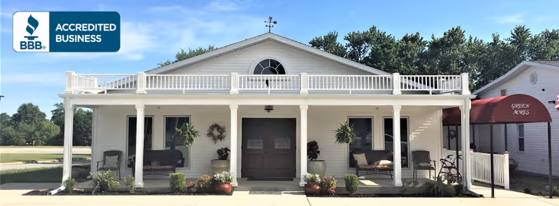 White single-story building with a covered porch, columns, potted plants, and a central double-door entrance under a balcony.