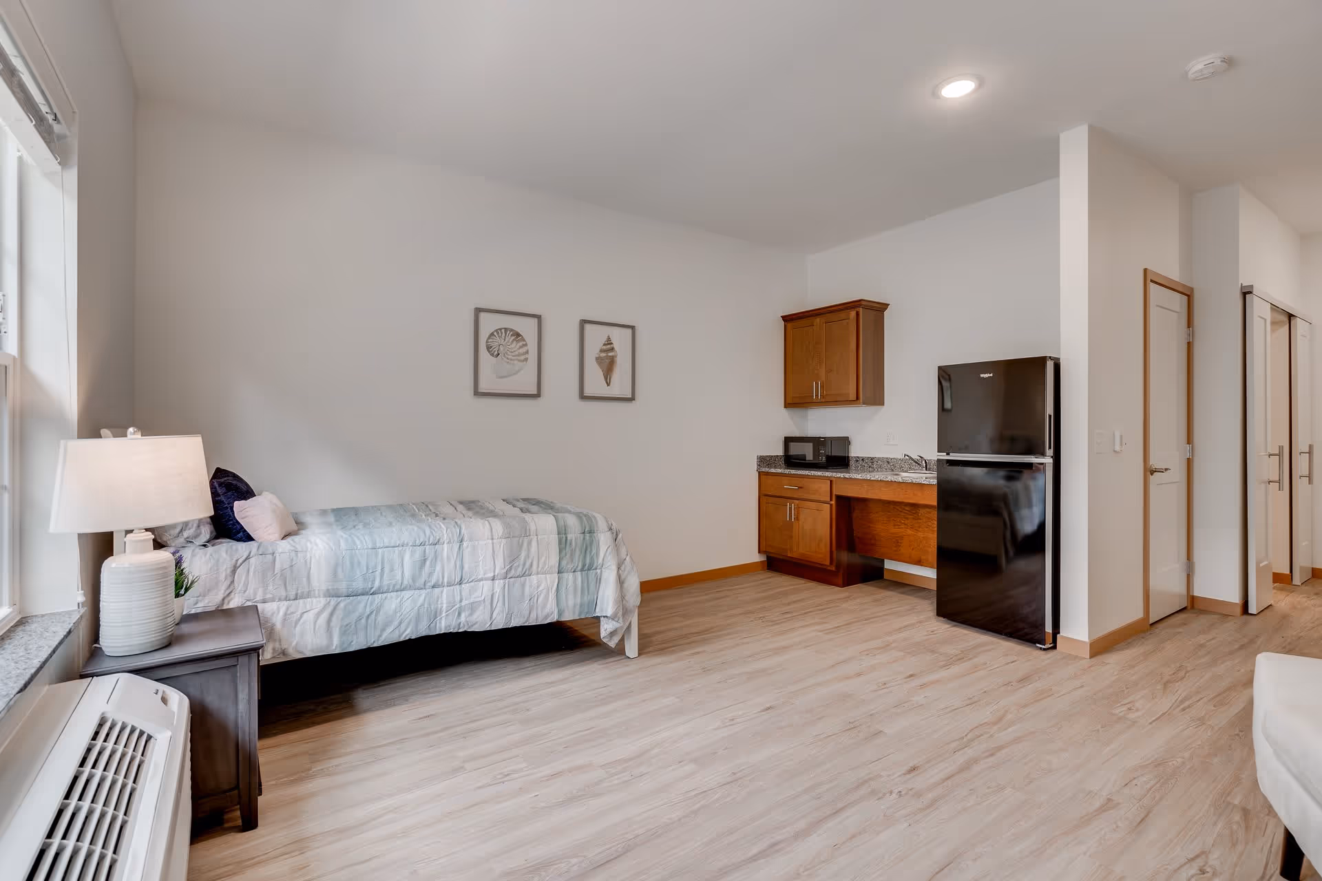 A bright and clean senior living studio apartment featuring a single bed with light blue bedding, a bedside table with a lamp and small plant, a kitchenette with wooden cabinets, a microwave, and a black refrigerator. The room has light wood flooring, white walls with two framed shell pictures, and a window letting in natural light.