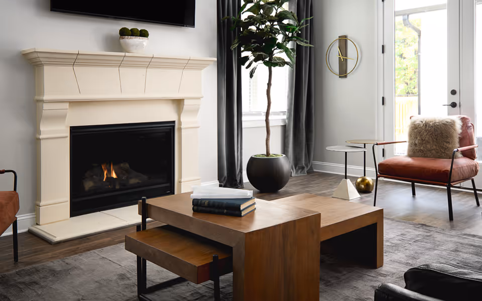A cozy living room with a lit fireplace, a wooden coffee table with books on it, a potted plant near a window with gray curtains, a modern wall clock, and two armchairs with cushions near a glass door leading outside.