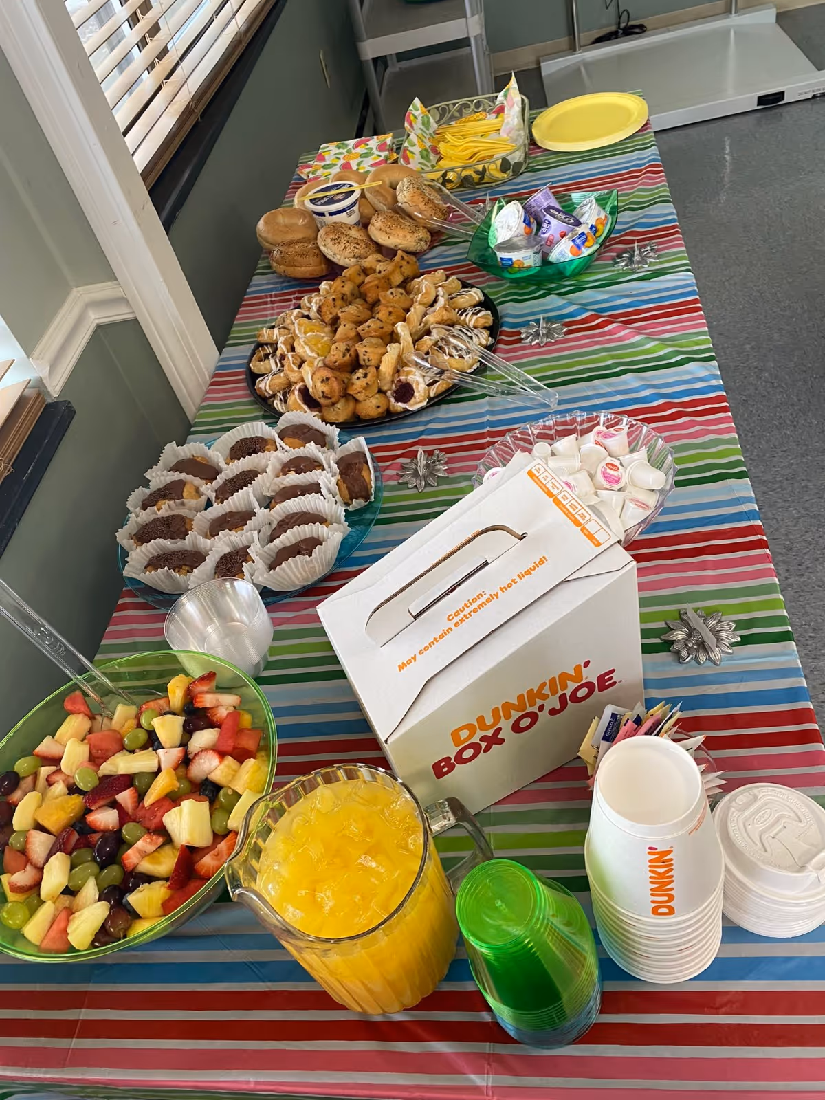 A table covered with a colorful striped tablecloth displaying a variety of breakfast items including a bowl of mixed fruit, muffins, bagels, pastries, a pitcher of orange juice, disposable cups, a Dunkin' Box O' Joe coffee box, and condiments such as creamers and sugar packets.