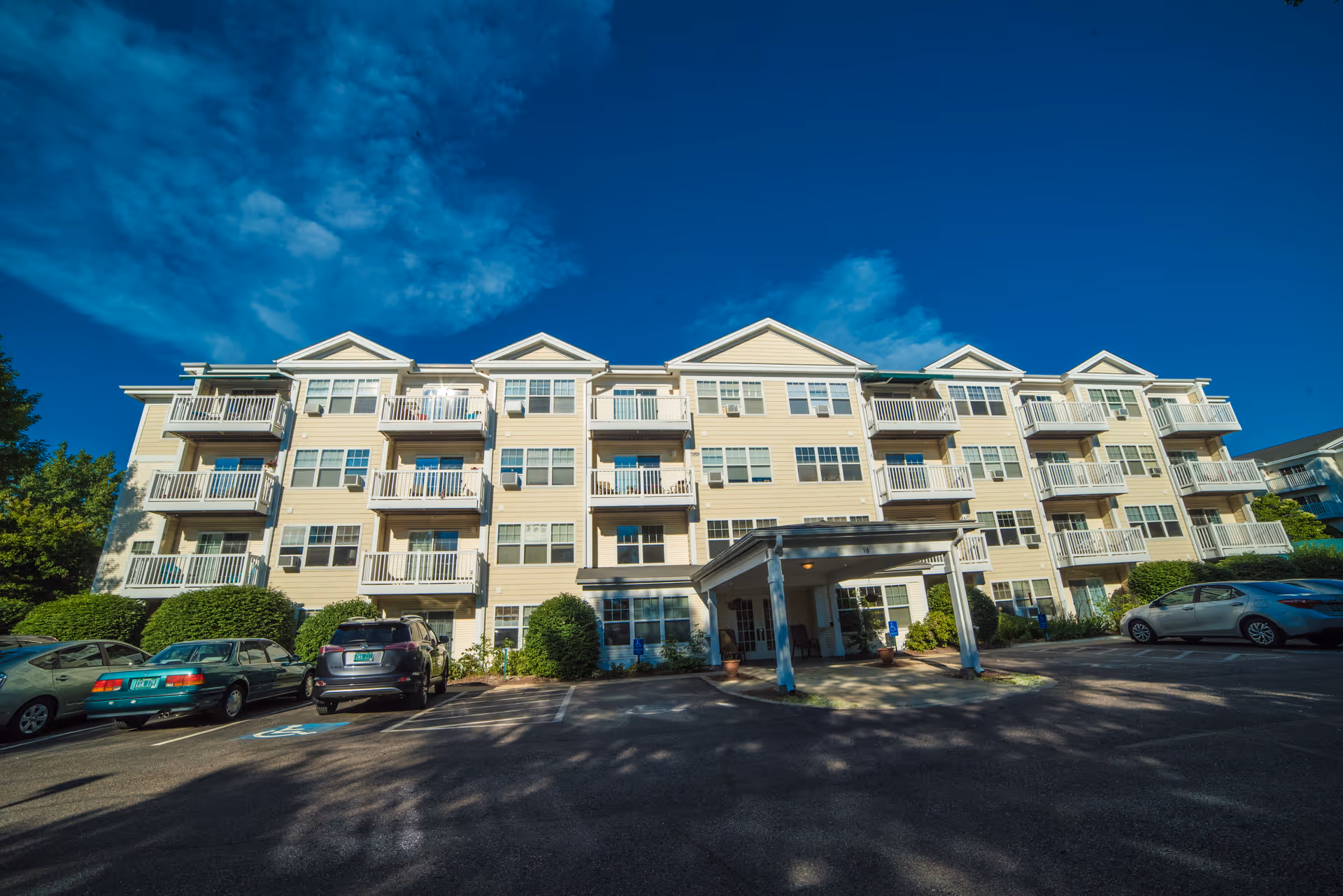 Exterior view of a multi-story senior living community building with balconies, a covered entrance, parked cars in front, and a bright blue sky with some clouds.