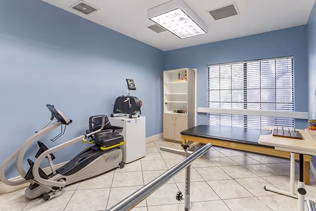 Rehabilitation/exercise room with a recumbent exercise bike, parallel bars, a treatment table, shelving cabinet, and a window with blinds.