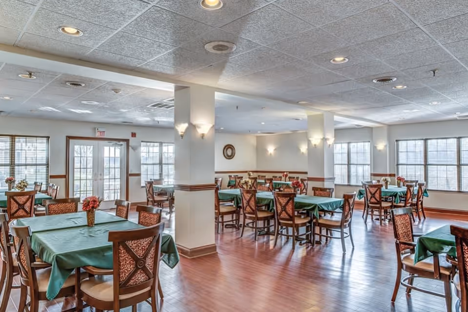 Bright communal dining room with several tables draped in green tablecloths and wooden chairs.