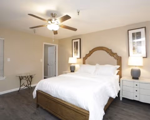 Well-lit bedroom with a wooden bed and headboard, white bedding, two nightstands with lamps, ceiling fan, and framed art.