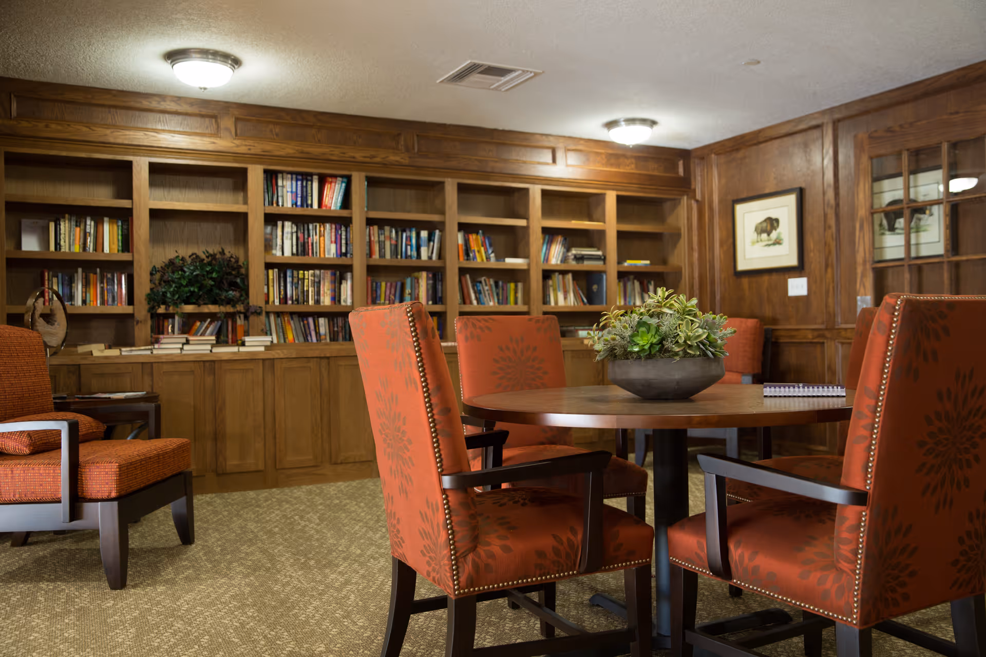 Cozy common room with a round wooden table surrounded by red upholstered chairs and built-in bookshelves along the back wall.