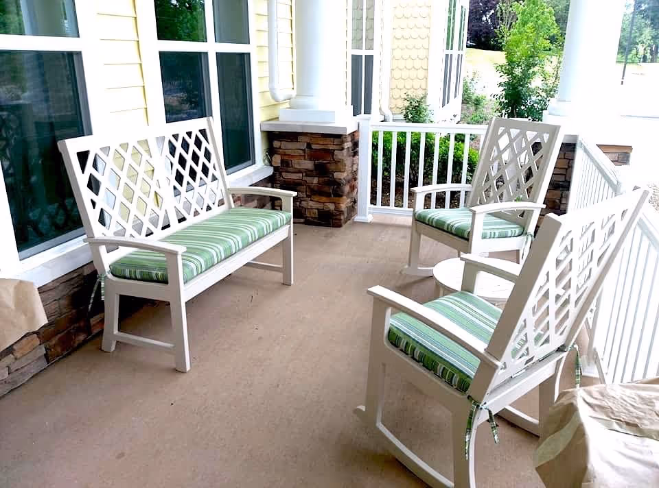A covered outdoor porch area with white wooden furniture including a bench and two rocking chairs with green and white striped cushions. The porch has white railings and stone accents on the columns, with windows and greenery visible in the background.