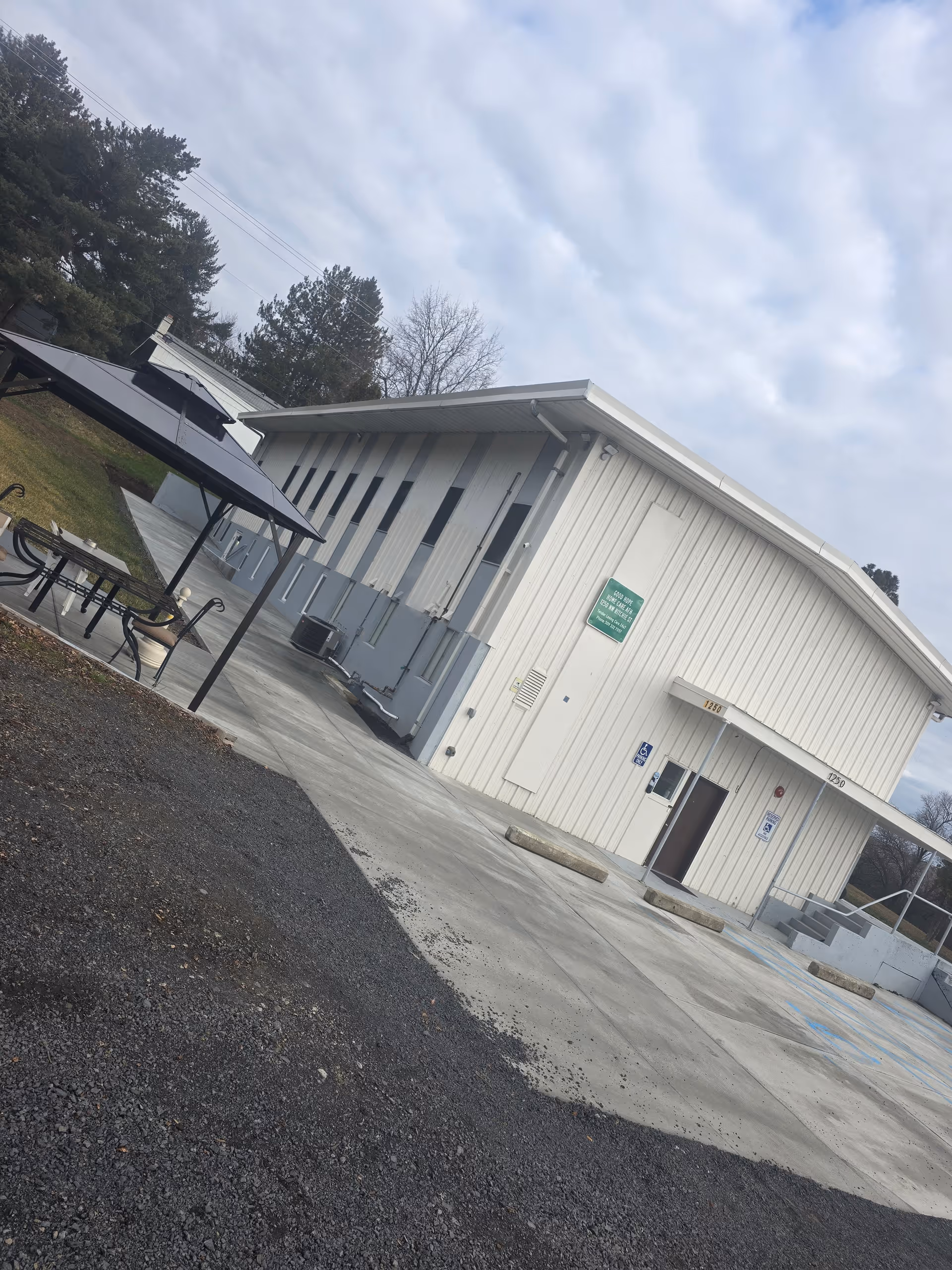 Exterior view of a white metal-sided care facility building with a paved parking area and a covered outdoor seating area.