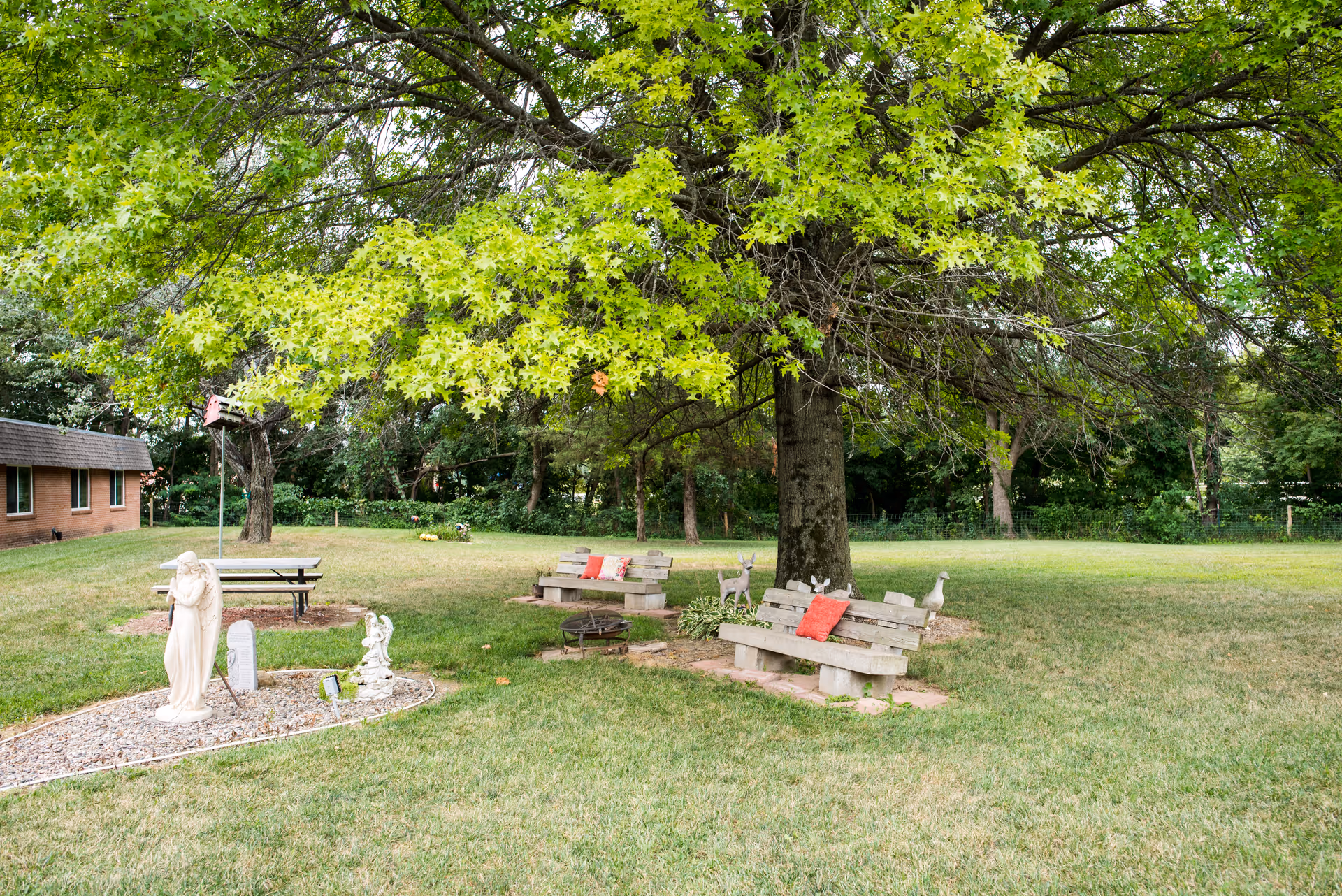 A peaceful outdoor garden area with a large leafy tree providing shade over two wooden benches with red cushions. There are small animal statues and a white angel statue on a gravel patch nearby. A brick building is visible on the left side, and the grassy lawn extends into the background with more trees.
