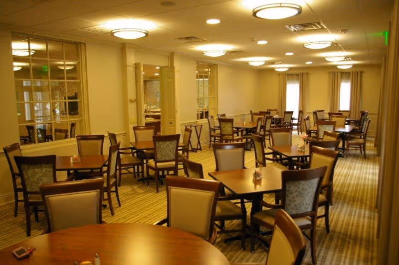 Large dining room with multiple wooden tables and upholstered chairs arranged in rows beneath round ceiling lights.
