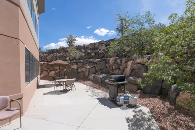 Outdoor patio area with a concrete floor, a table with an umbrella and chairs, a barbecue grill with propane tanks, surrounded by a rocky retaining wall and some trees under a blue sky with a few clouds.