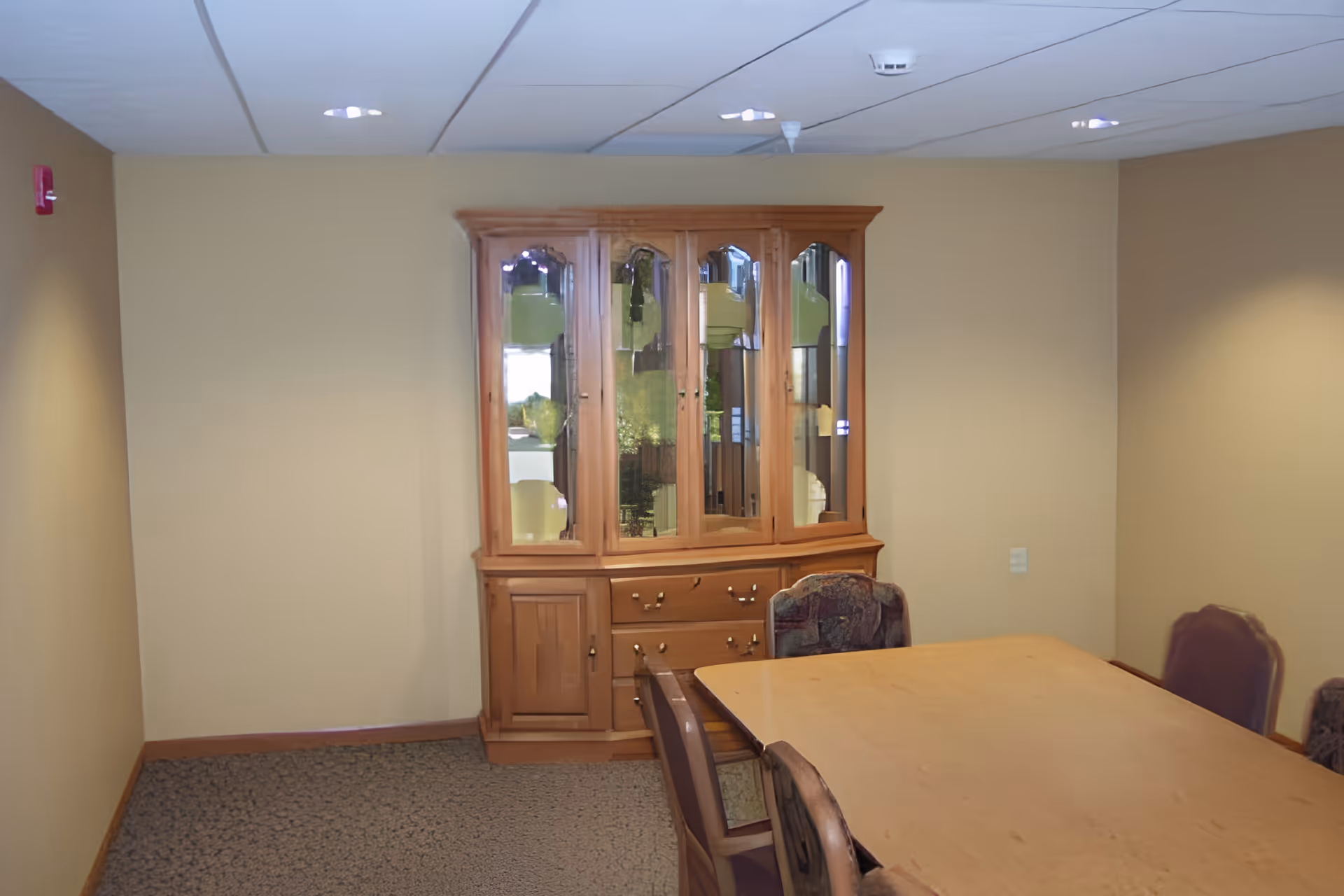 A small dining room with a wooden dining table surrounded by chairs and a wooden china cabinet with glass doors against a beige wall.