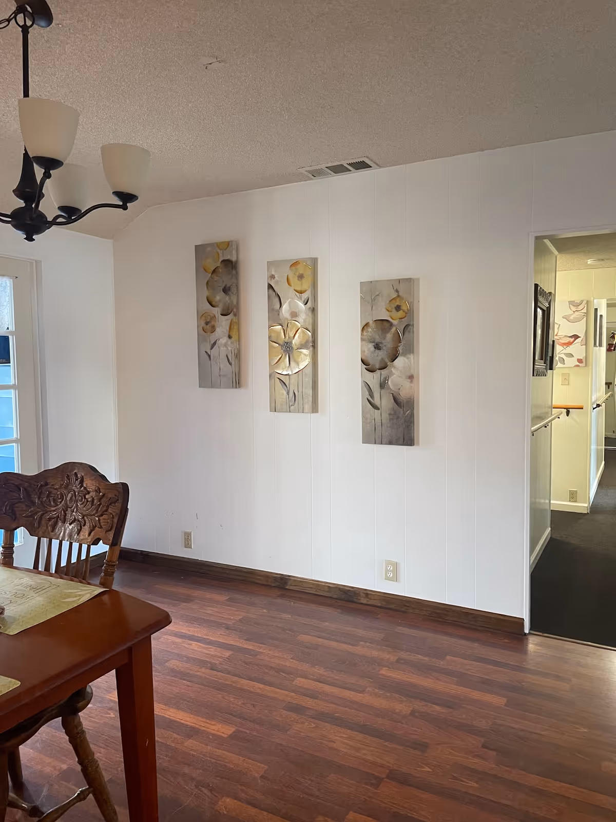 Interior view of a dining area with wooden flooring, a wooden dining table and chair, a chandelier with three lights, and three vertical floral paintings on a white wall. There is a doorway leading to a hallway with handrails and additional artwork visible.