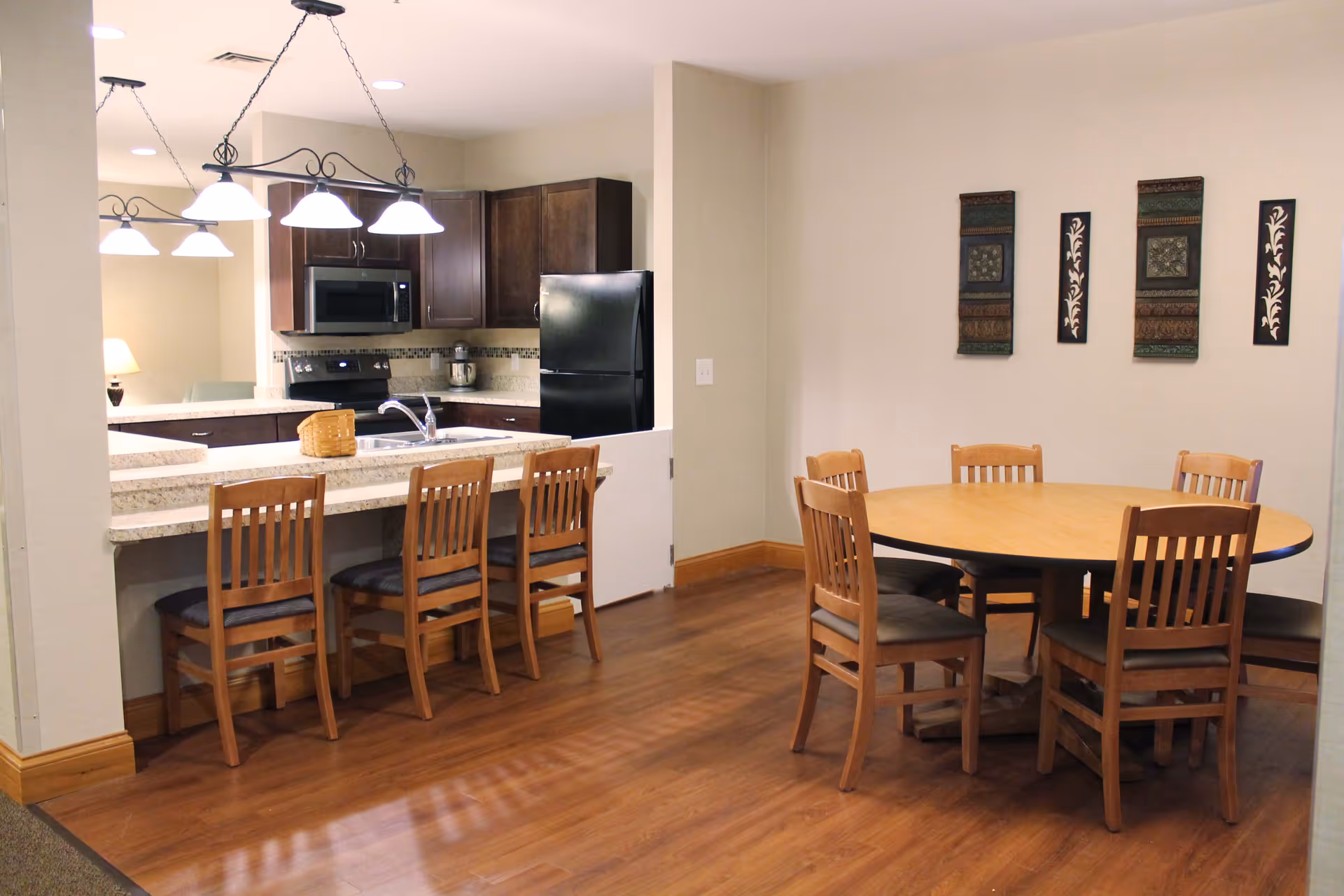Interior view of a kitchen and dining area in a senior living facility. The kitchen features dark wood cabinets, a black refrigerator, a microwave, and a stove. There is a countertop with a sink and four wooden chairs lined up along the counter. Adjacent to the kitchen is a round wooden dining table with six matching wooden chairs. The walls are light-colored with decorative wall hangings, and the floor is a warm-toned wood.