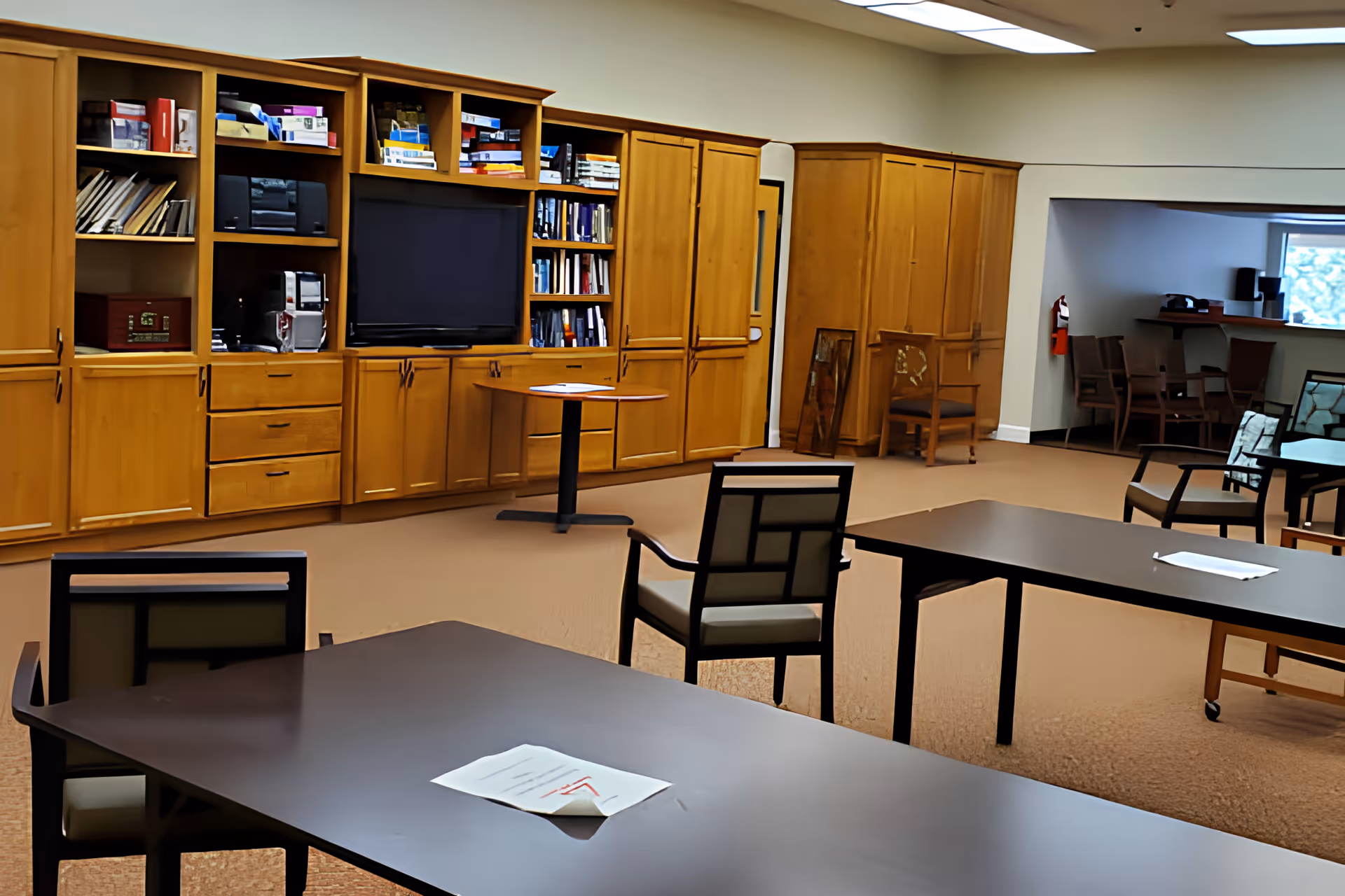 A spacious common room with multiple tables and chairs arranged for seating. Along one wall is a large wooden built-in shelving unit with cabinets, books, a television, and various items. In the background, there is a small seating area with additional chairs and a window.