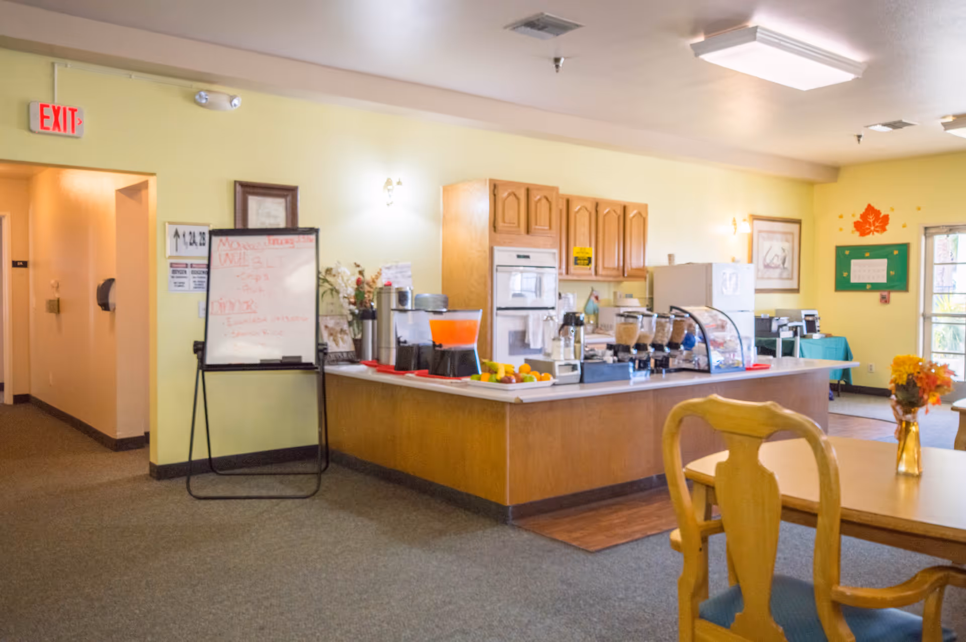 Interior view of a communal kitchen and dining area in an assisted living community. The kitchen features wooden cabinets, a built-in oven, a refrigerator, and a counter with beverage dispensers, cereal containers, and fruit. A whiteboard with daily menu items is visible near an exit sign. A wooden dining table with chairs and a small vase with flowers is in the foreground. The walls are painted light yellow, and there are framed pictures and decorations on the walls.