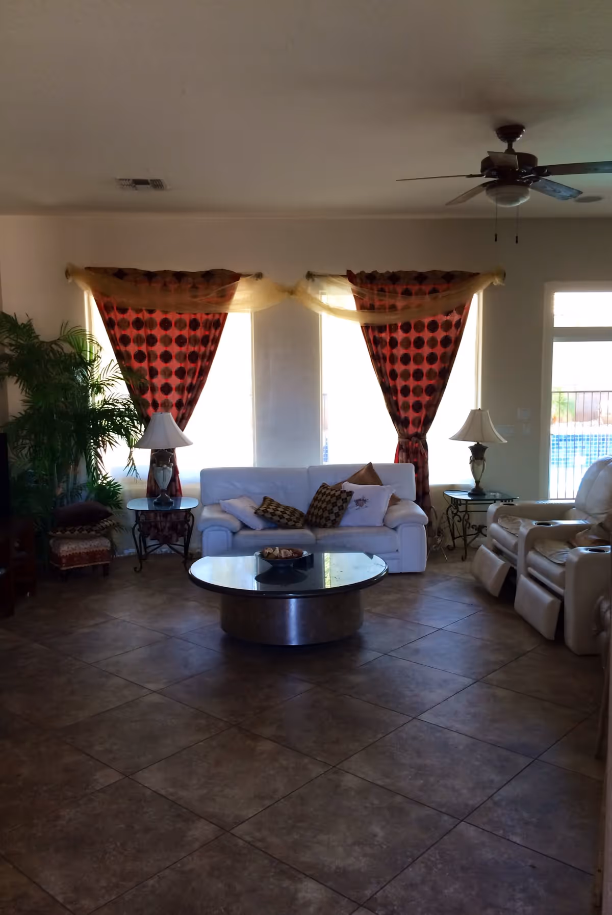 A living room with a white sofa adorned with multiple cushions, two side tables each holding a lamp, a round coffee table in the center, red and black patterned curtains with a sheer yellow swag draped over the top, a ceiling fan, and a large potted plant on the left side. There are also two beige recliner chairs on the right side near a window showing an outdoor pool area.